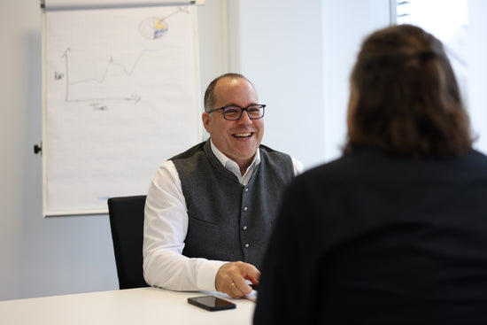 Manuel Weiskopf lacht im persönlichen Beratungsgespräch; im Hintergrund Flipchart mit Fortschrittsgrafik.